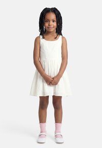 Jeune fille aux cheveux tressés portant une robe blanche sans manches, des chaussettes roses et des chaussures Mary Jane blanches.