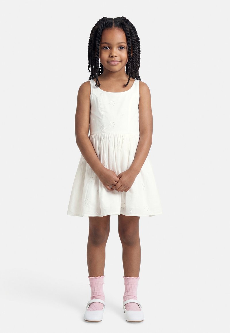 Jeune fille aux cheveux tressés portant une robe blanche sans manches, des chaussettes roses et des chaussures Mary Jane blanches.