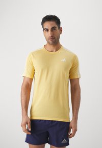 Camiseta atlética amarilla de manga corta, con cuello redondo y logotipo de Adidas en blanco. Presenta tres stripes blancas en los hombros. Tela suave, ajuste regular.