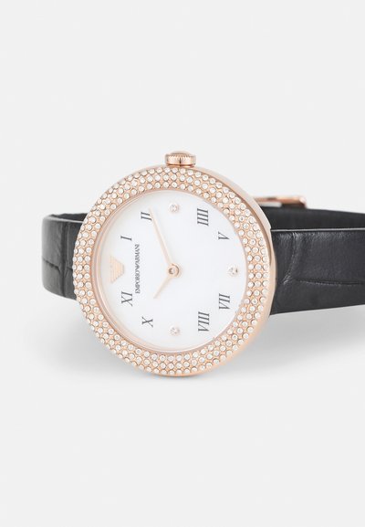 Montre ronde avec un cadran blanc, des index en chiffres romains, une lunette en rose gold ornée de cristaux, et un bracelet en cuir noir.