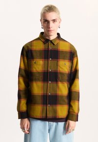 Camicia in flanella a quadri gialli e neri con maniche lunghe, due tasche sul petto e colletto con bottoni. Tessuto liscio, vestibilità casual e bottoni in metallo.