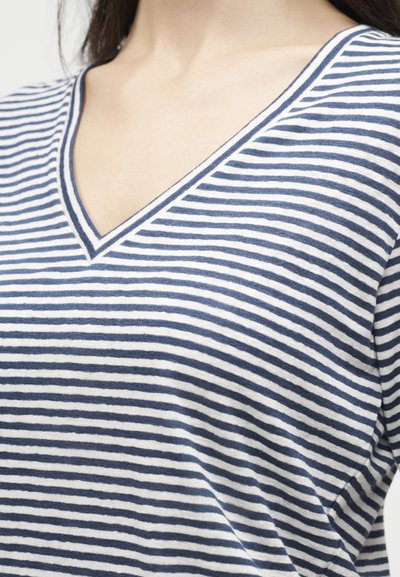 Γυναίκα που φοράει μπλουζάκι με μπλε και άσπρες οριζόντιες ρίγες και V-λαιμόκοψη, εμφανίζοντας το πάνω μέρος του στήθους και των ώμων.