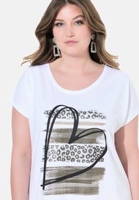 Vit kortärmad t-shirt med ett hjärtdesign i leopardmönster och metalliska detaljer i guld- och bronsränder. Mjuk bomullstyg.