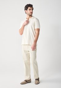 Hellbeige gerippte Poloshirt, cremefarbene Hose und gemusterte braune Slipper. Kurze Ärmel, ein Kragen und eine lockere Passform.