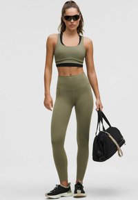 Olíva zöld sport melltartóban és leggingsben, fekete sportcipőben, napszemüvegben áll, miközben egy fekete tornazsákot tart, egy sima háttér előtt.