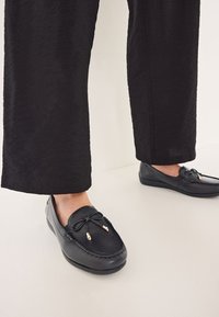 Schwarze Lederloafers mit einer strukturierten Oberfläche, verziert mit einer dekorativen Schleife und goldenen Akzenten an den Schnürsenkeln; kombiniert mit lockeren schwarzen Hosen.