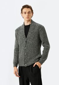 Grauer Strickcardigan mit Schalkragen, ausgestattet mit fünf schwarzen Knöpfen, gerippter Textur und langen Ärmeln. Getragen über einem schwarzen T-Shirt.