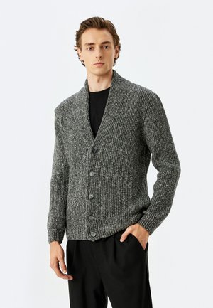 Cárdigan de punto gris con cuello de chal, que presenta cinco botones negros, textura de canalé y mangas largas. Lleva puesto sobre una camiseta negra.