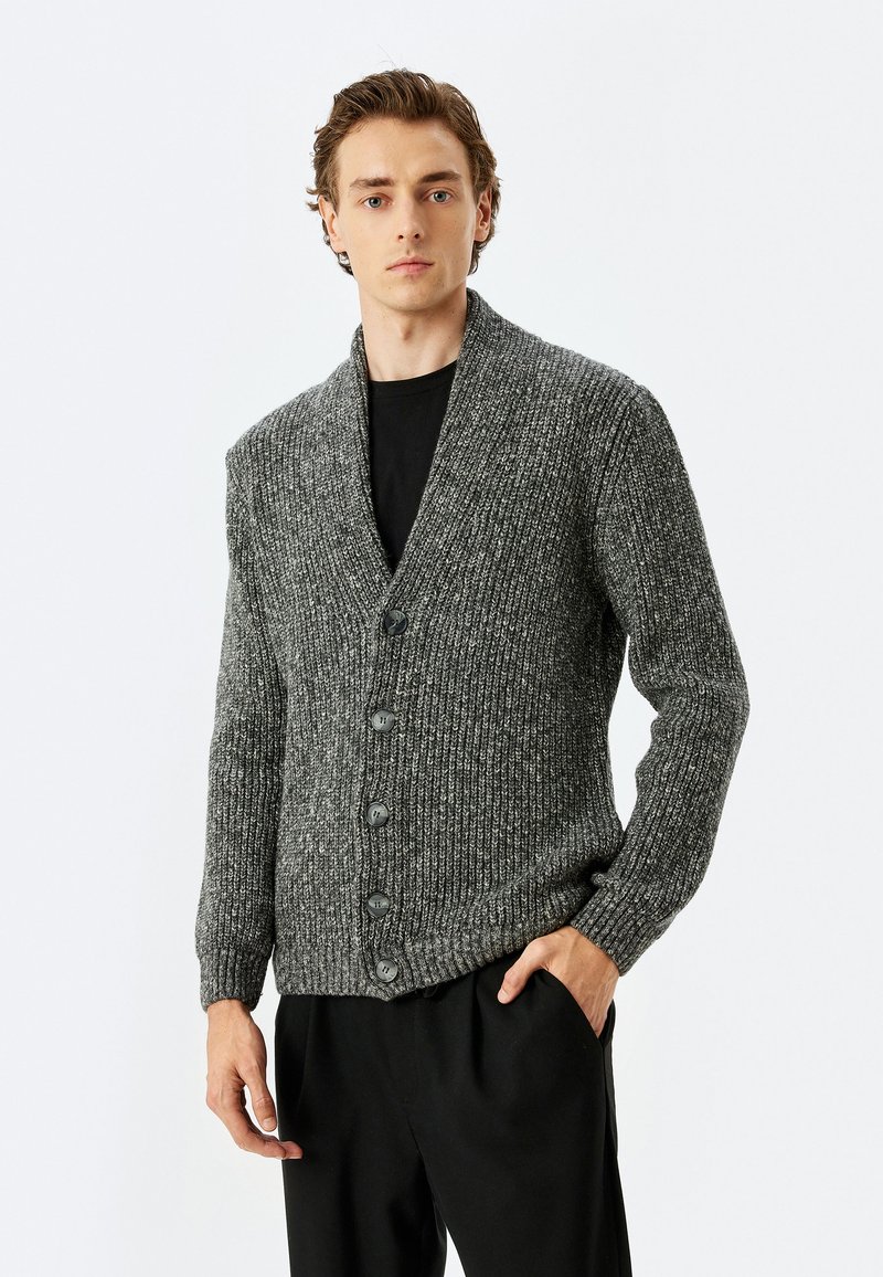 Grauer Strickcardigan mit Schalkragen, ausgestattet mit fünf schwarzen Knöpfen, gerippter Textur und langen Ärmeln. Getragen über einem schwarzen T-Shirt.