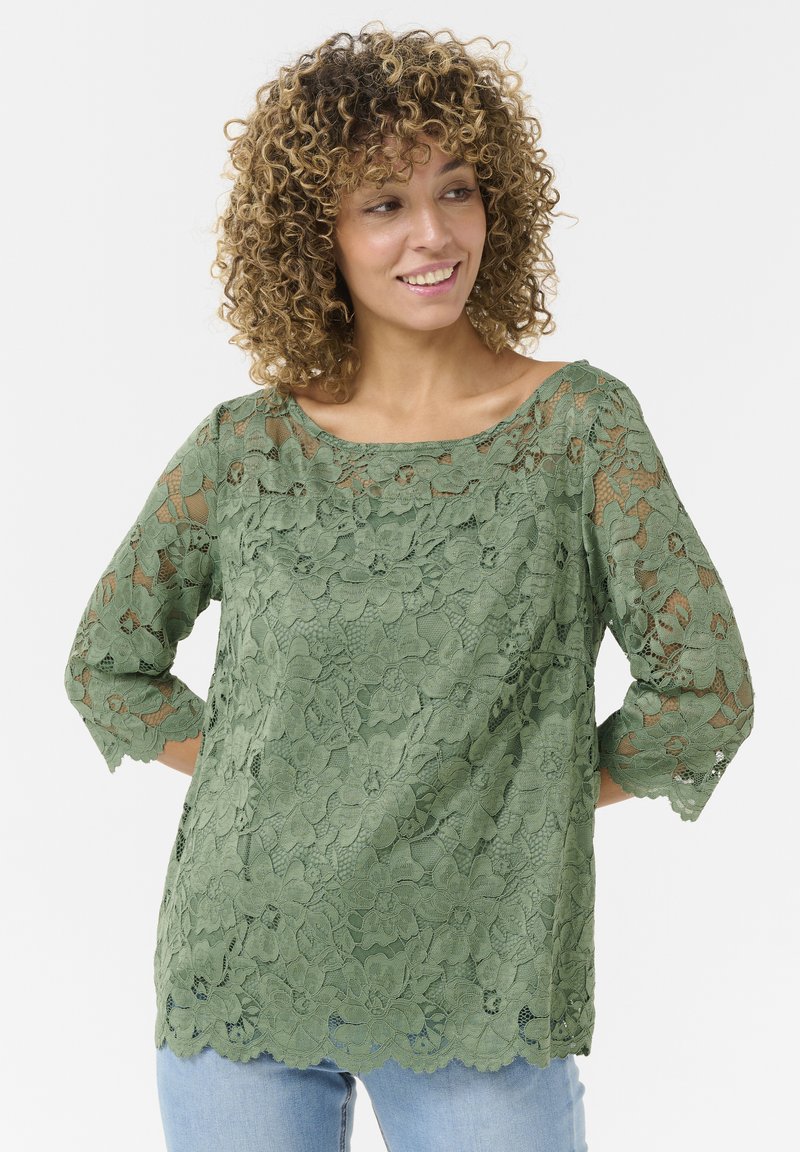 Top de encaje verde con mangas de tres cuartos, patrón floral, dobladillo festoneado y detalles transparentes. Combinado con un denim azul claro.