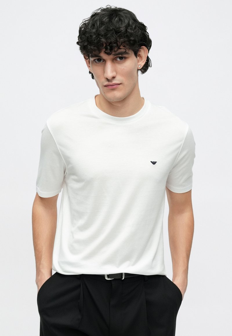 T-shirt bianca a maniche corte realizzata in tessuto morbido, con un piccolo logo nero sul petto e una vestibilità comoda. Indossata con pantaloni neri.