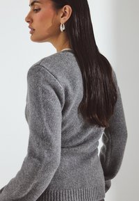 Grau gestrickter Pullover mit langen Ärmeln, geripptem Saum und taillierter Silhouette. Die Textur wirkt weich und warm und ergänzt das schlanke Design.