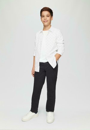 Weißes Hemd mit kleinen schwarzen Mustern, über einem weißen T-Shirt getragen, kombiniert mit schwarzen Hosen und weißen Sneakern.