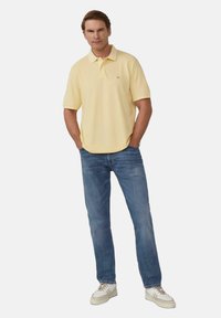 Żółta koszulka polo z kołnierzykiem, zestawiona z jasnoniebieskimi dżinsami. Model stoi z rękami na biodrach, nosząc białe i beżowe sneakersy.