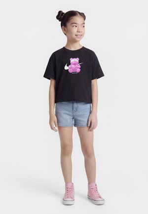 Ragazza giovane con pettinatura a doppio chignon, indossa una t-shirt nera con grafica di orso rosa, pantaloncini di jeans e sneakers alte rosa, in piedi e guarda verso destra.