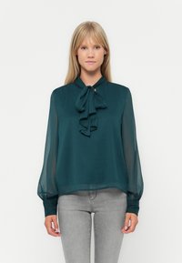 Τιρκουάζ μπλούζα με φιόγκο στον λαιμό, ελαφρύ διάφανο ύφασμα, μακριά μανίκια με ελαστικούς καρπούς και χαλαρή εφαρμογή. Συνδυάζεται με γκρι τζιν.