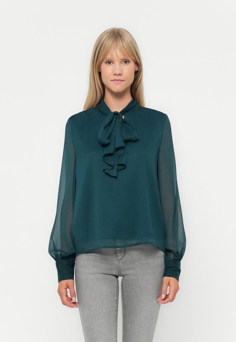 Τιρκουάζ μπλούζα με φιόγκο στον λαιμό, ελαφρύ διάφανο ύφασμα, μακριά μανίκια με ελαστικούς καρπούς και χαλαρή εφαρμογή. Συνδυάζεται με γκρι τζιν.