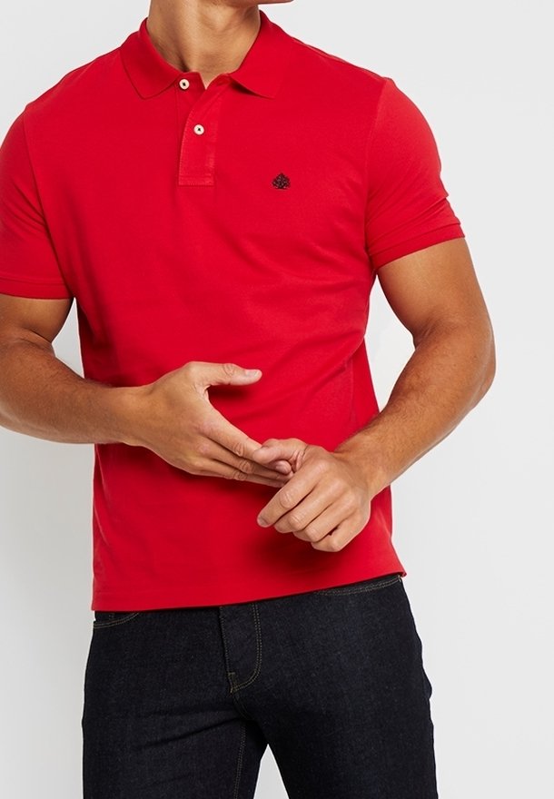 Homme portant un polo rouge à manches courtes avec des boutons et un jean noir, ajustant ses doigts avec les mains devant le torse.
