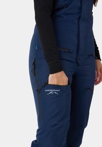 Marineblauwe geïsoleerde overall met een ritszak, logo-detail, verstelbare banden en ademend materiaal voor buitengebruik.