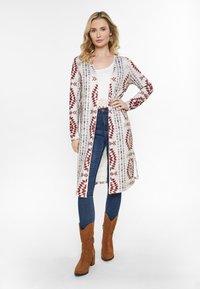 Langer Cardigan in Creme mit roten geometrischen Mustern, offene Front, über einem weißen Oberteil und dunklen Jeans getragen, ergänzt durch braune Ankle Boots.