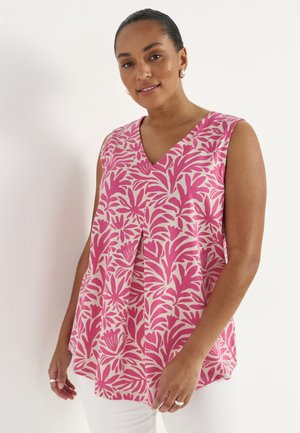 Kvinde iført ærmeløs pink og hvid blomstret bluse med V-udskæring og hvide bukser, stående foran en ensfarvet lys baggrund.