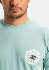 Světle modré bavlněné tričko s kulatým logem, na kterém je napsáno "Quiksilver" v bílém a tmavém písmu. Klasický design s kulatým výstřihem, hladká textura.