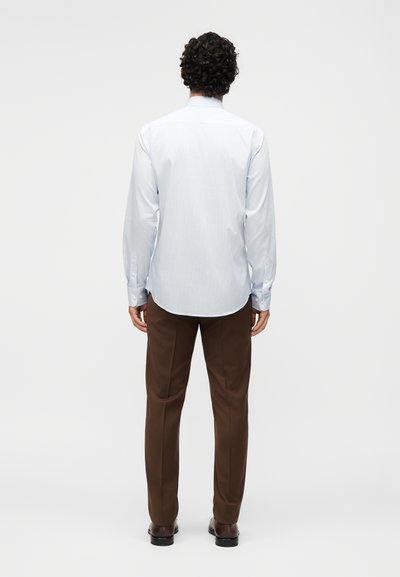 Άνδρας με σγουρά μαλλιά, φοράει ένα ανοιχτό μπλε ριγέ πουκάμισο, καφέ παντελόνια και σκούρα παπούτσια, στέκεται με την πλάτη γυρισμένη σε απλό φόντο.