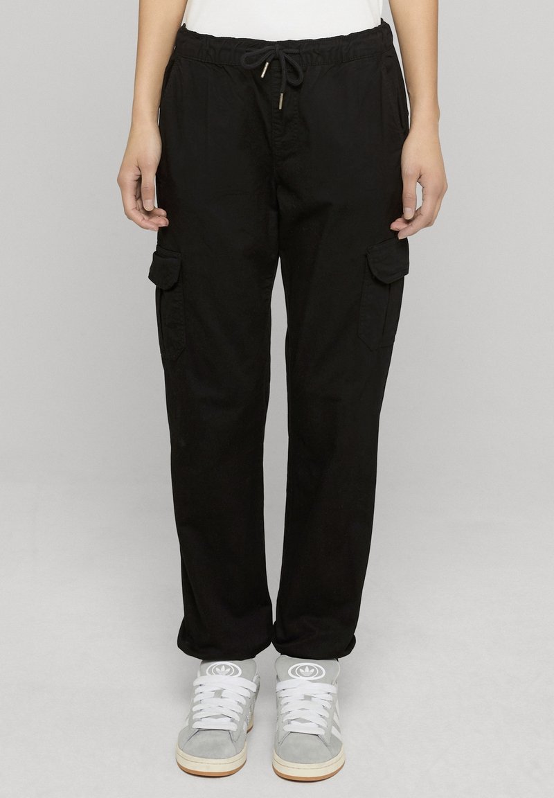 Personne portant un pantalon cargo noir avec des poches avant et des baskets blanches avec des accents gris, debout devant un fond gris clair uni.