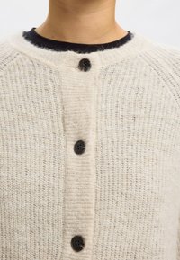 Jasnobeżowy, dziergany kardigan z miękkiej włóczki, z okrągłym dekoltem i sześcioma czarnymi plastikowymi guzikami z przodu.