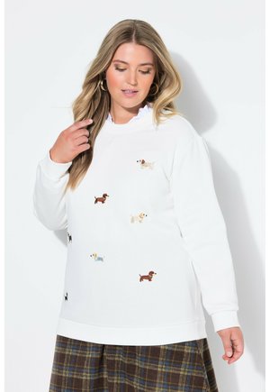 Kvinde iført hvid sweatshirt dekoreret med små farverige hundeillustrationer og et ternet skørt, poserende mod en hvid baggrund.
