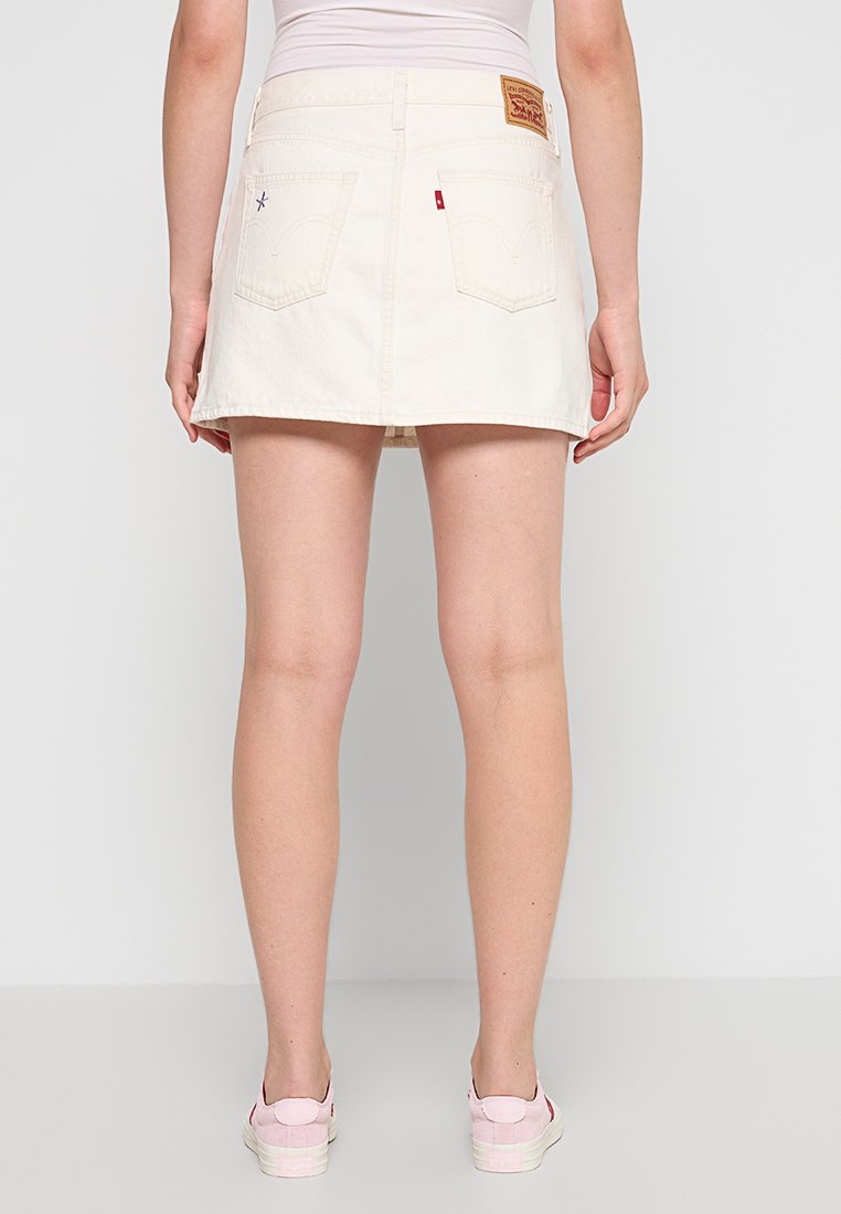 Jupe mini en denim blanc avec une taille ajustée, deux poches arrière et une étiquette en cuir de marque. Les coutures visibles ajoutent de la texture.