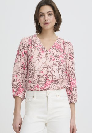 Kobieta ubrana w różową bluzkę w kwiaty z dekoltem w serek i rękawami do połowy przedramienia oraz białe spodnie z wysokim stanem, stojąca na jednolitym tle.