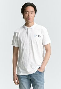 Weißes Poloshirt mit strukturiertem Finish, versehen mit einem hellblauen "HAVANA TIMES" Grafik, kombiniert mit hellblauen Jeans.