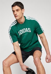 Zöld pamut póló fehér Adidas logóval, fehér csíkokkal a trikón. Modell fekete sortot visel, és egy kettlebellt tart.