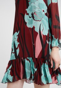 Robe fleurie avec de grandes fleurs bleues et roses, tiges noires sur tissu rouge foncé, manches longues transparentes avec poignets à volants et ourlet à volants.