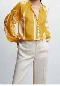 Gelbe Bluse mit weißen Mustern, Knopfleiste und lässiger Passform. Kombiniert mit cremefarbener, maßgeschneiderter Hose.