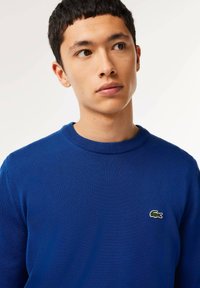 Maglione blu lavorato a maglia con scollatura rotonda e un piccolo logo di coccodrillo verde sul lato sinistro del petto, caratterizzato da una texture liscia e un design aderente.