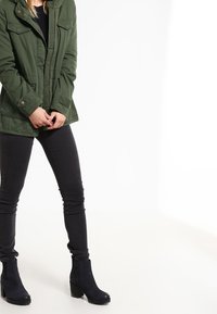 Parka de color verde oliva con bolsillos abotonados, llevada sobre una camiseta negra y unos vaqueros pitillo oscuros. Botines negros con tacón bloque.