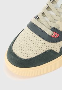 Sneaker avec une tige en cuir beige perforé, des accents en daim vert foncé, des lacets gris clair et une semelle en caoutchouc crème. Équipée d'un détail matériel rouge.