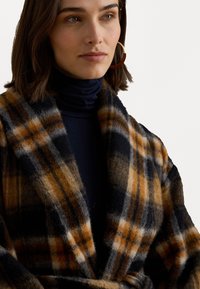 Cappotto di lana a quadri con colori arancione, grigio e nero; texture morbida, collo ampio e design aperto frontalmente. Strato interno liscio visibile.
