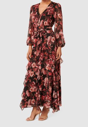Femme portant une longue robe noire avec des motifs floraux rouges et roses, des manches longues transparentes, un décolleté en V et des talons beiges à bouts ouverts.