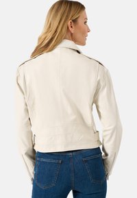 Cremfarbene Lederjacke mit tailliertem Design, ausgestattet mit Schulterklappen, einem taillierten Bund und Reißverschlussan den Ärmelabschlüssen. Über dunkelhimmelblauen Jeans getragen.