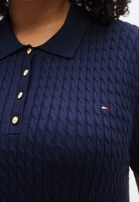 Πλεκτό μπλε μπλουζάκι πόλο σε ναυτικό μπλε χρώμα με υφή καλωδιακού μοτίβου, πέντε χρυσά κουμπιά και ένα μικρό κόκκινο, λευκό και μπλε λογότυπο στο στήθος.