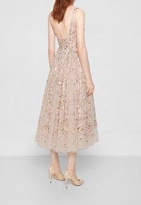 Mujer vestida con un vestido midi beige sin mangas y sin espalda, con bordados florales, combinado con zapatos de tacón nude, de pie sobre un fondo neutro.