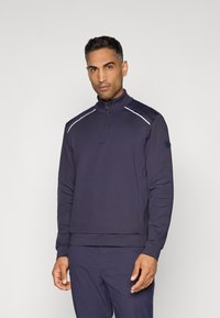 Sweatshirt bleu marine à zip 1/4 en tissu lisse, avec des piping blancs contrastants, un col haut et un patch logo sur la manche gauche.