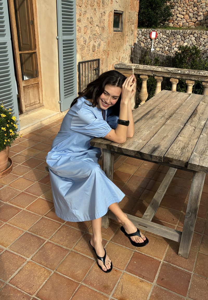 Femme en robe bleu clair assise sur le sol de la terrasse, appuyée sur une table en bois, souriante avec les mains posées sur la tête dans un cadre extérieur ensoleillé.