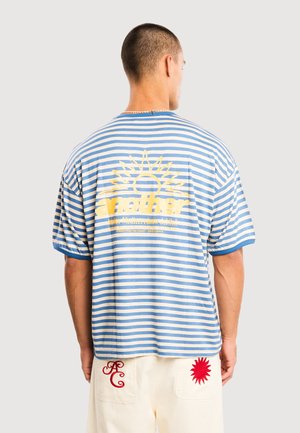 Mand iført blå- og hvidstribet T-shirt med gul solgrafik og tekst samt cremefarvede shorts med rød sol og bogstaver på lommerne.