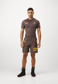 Roupa desportiva da UFC com uma camisola ajustada de mangas curtas castanha com detalhes em amarelo e calções castanhos combinando com um logo amarelo ousado e padrão.