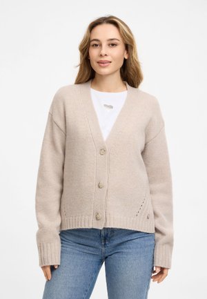 Junge Frau mit hellbraunen Haaren trägt einen beigen, geknöpften Cardigan über weißem Top und blauen Jeans, steht vor einem schlichten weißen Hintergrund.