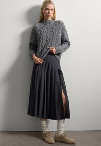 Femme portant un pull en tricot gris à motifs torsadés, une jupe midi plissée bleu marine avec une fente sur le côté, des chaussettes blanches froissées et des baskets beiges se tenant contre un mur blanc.